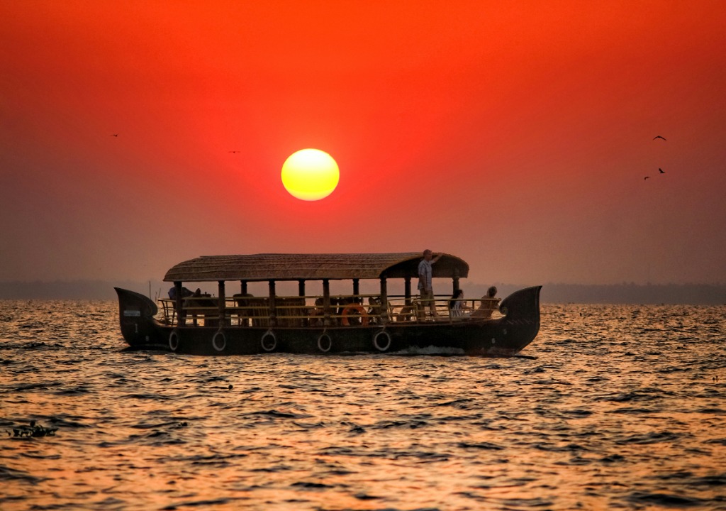 A traditional wooden houseboat sailing on calm Vembanad lake at sunset in Kerala